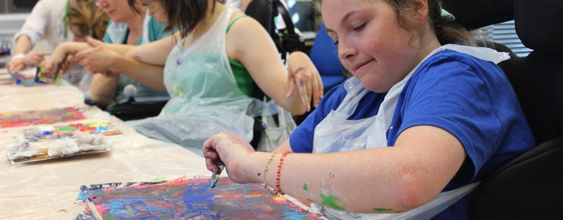 Treloar College students at a long desk in the art classroom, working on their painting by using credit cards to place paint. All look very focused on the task.