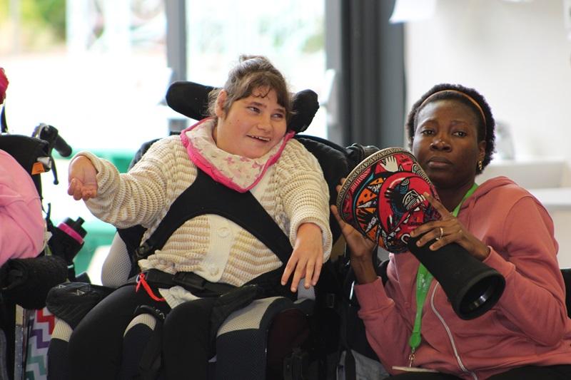 Treloar College student smiling and tapping a drum being held by her assistant during African drumming workshops.