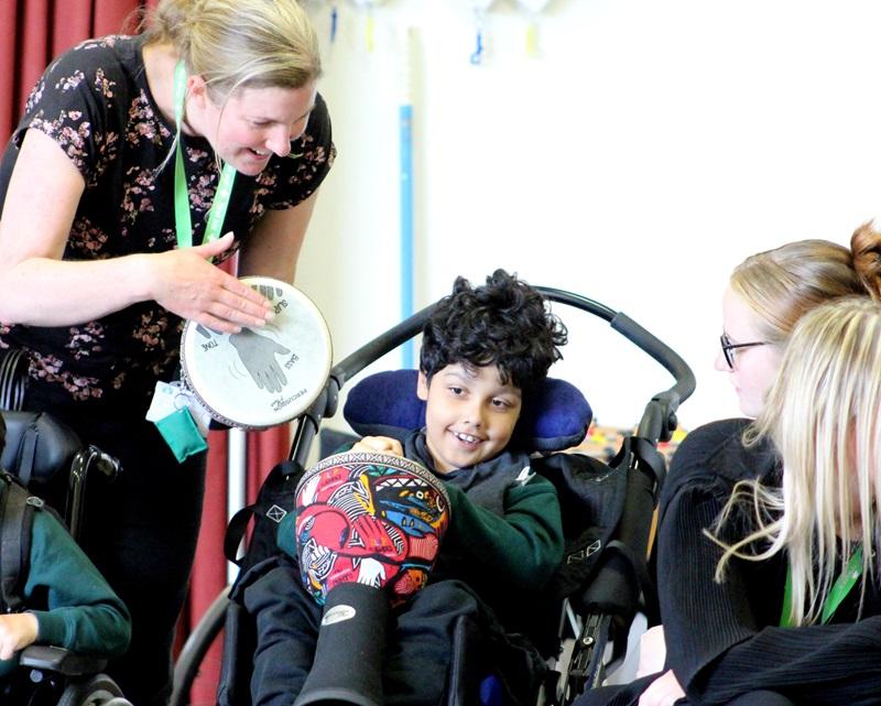 Treloar School student holding a drum during African drumming workshops; his teacher is leaning over him and holding her own drum - all having a good time.