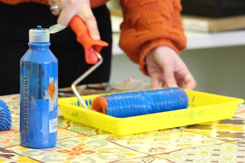 A yellow tray with blue paint in and a bottle of blue paint next to it: Rachel the art therapist is using a roller to dip in the paint.