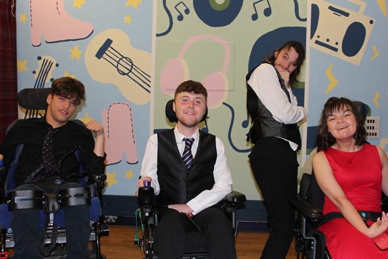 4 Treloar College students at their prom, all dressed up, one of them is standing; all smiling at the camera.