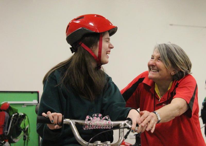 Treloar's student while triking, holding her both hands on the handlebars, wearing shiny red helmet, looking at the instructor who is helping her control the trike. Both smiling at each other.