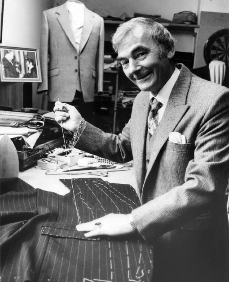 Black and white photo of Peter Little: he is sawing a suit, holding a thread and smiling at the camera.