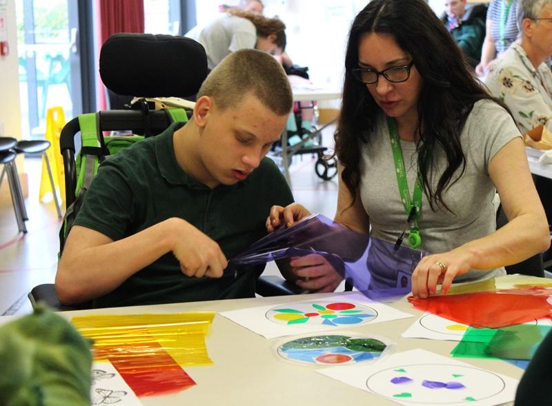 Treloar's student using scissors to cut his stained glass design sheet which is purple; his assistant is helping him.