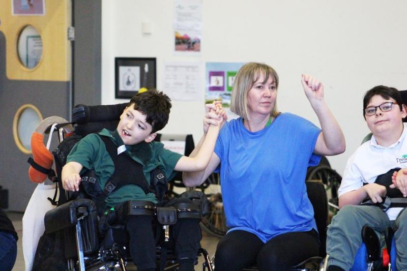 2 Treloar's students taking part in a dance exercise; student support assistant is holding her hand with one of the boys and moving to the sound of music.