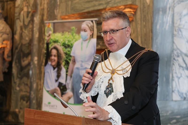 Portrait of Alastair King who is standing at a podium and holding a microphone in his right hand.