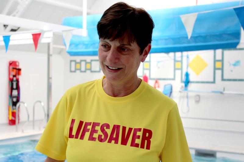 Kim Smith, Sports Coach and Event Leader at Treloar's pictured wearing bright yellow T-shirt with a red writing on her chest saying Lifesaver; in the background, there's a swimming pool area.