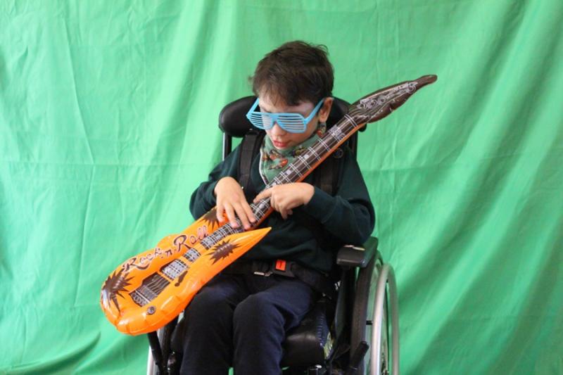 Treloar School student posing for a photo holding a guitar and wearing blue plastic sunglasses; behind him, there is a greenscreen.