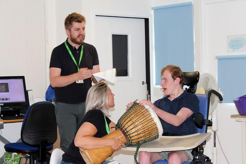 Tom Clarke in the classroom teaching his class: he is standing in front of one student who is playing a big drum - the drum is being held by his assistant.