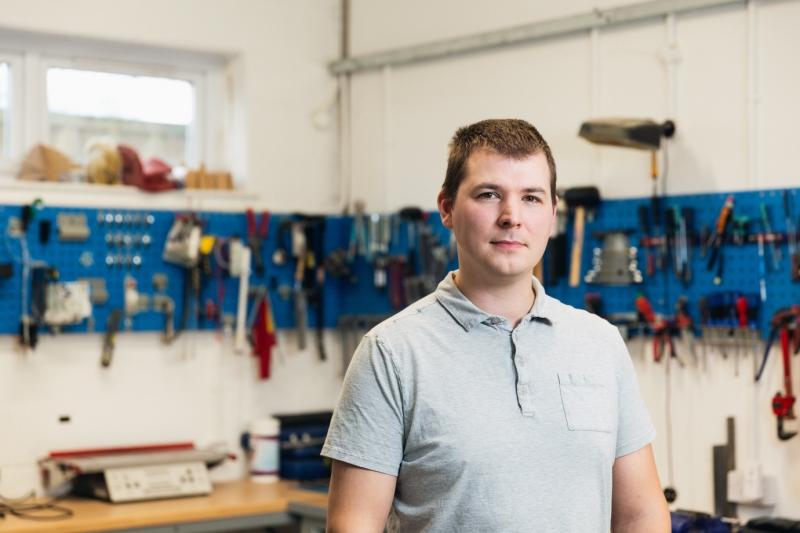 Portrait of Dean Hall, an Assistive Technologist at Treloar’s. In the background, multiple tools hanging on the walls of the workshop. 