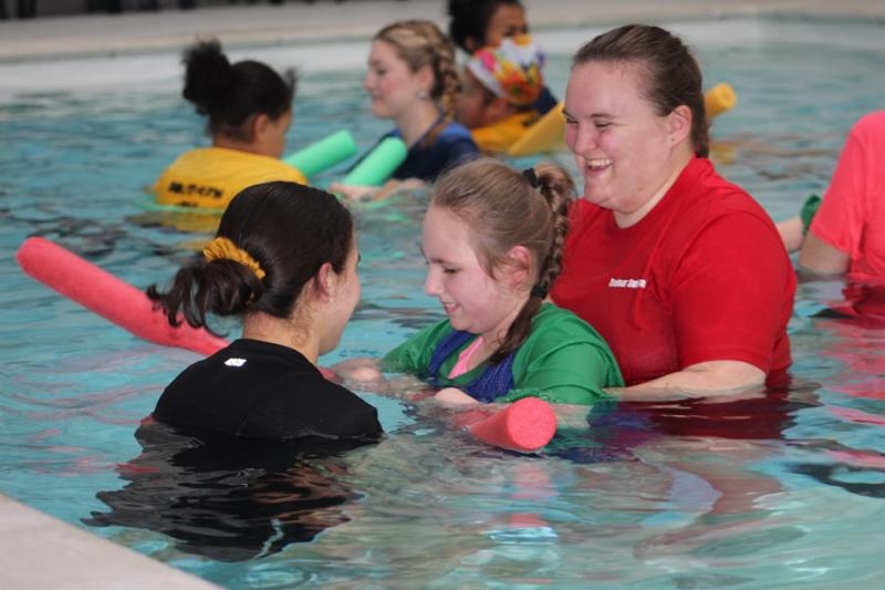 Treloar School student in the pool, supported by 2 members of staff holding onto a pink swimming noodle; in the background other students and staff.