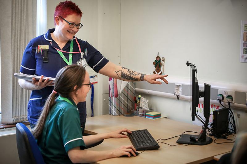 Treloar's nurse and medicine management technician in an office; the technician is sitting at her desk looking at a PC screen while the nurse is standing right next to her and pointing at the desktop, explaining something to the technician.