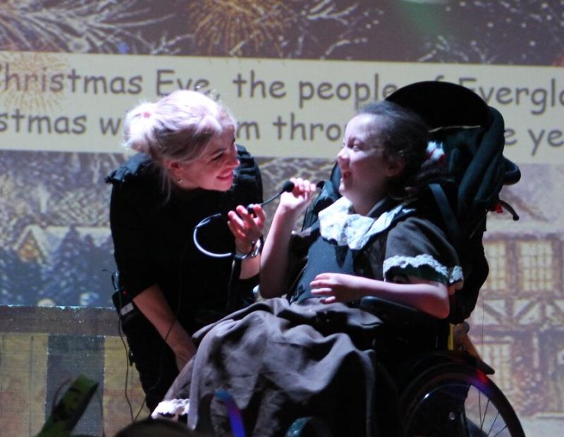 Donna, Deputy Lead Student Support Assistant at Treloar’s, on the stage with Treloar School student during a Christmas performance: she is holding a microphone helping student sing; both are smiling and having a good time.