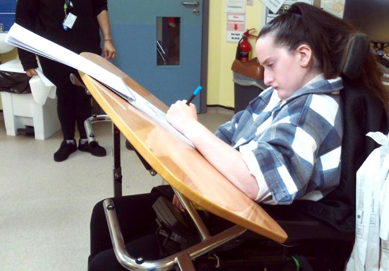 Treloar College student using a sloping desk that enables her to make notes in her book much more easily. She is in a classroom setting, she looks focused on her task.