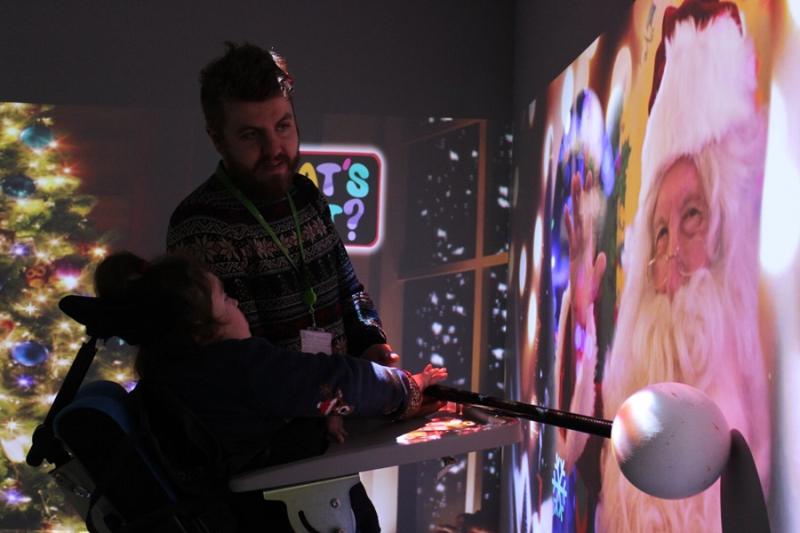 An interactive projections on the wall of Santa, accompanied by scents and sounds: Treloar College student is being assisted by her teacher to interactive with the projection by holding a big pole with a round end.