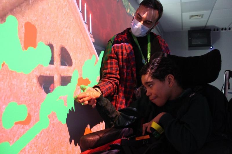 An interactive projections on the wall of a gingerbread house, accompanied by scents and sounds: Treloar School student is being assisted by his teacher to interactive with the projection by touching the wall.