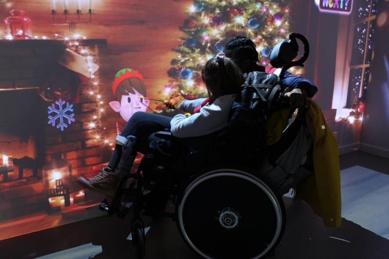 An interactive projection on the wall of a Christmas elf, accompanied by scents and sounds: Treloar College student is interacting with the projection by using a stick - her assistant is helping the student hold the stick.