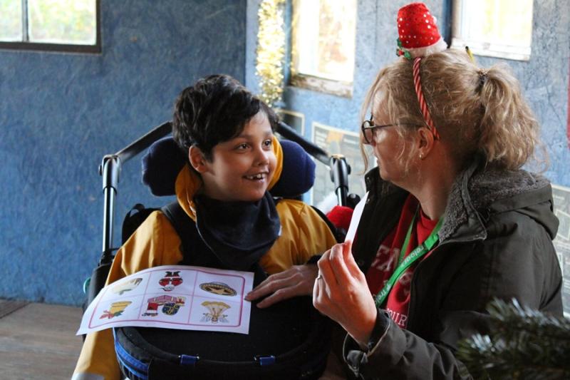 Treloar's primary student playing Christmas bingo with his assistant: the bingo sheet is on his lap and his assistant is showing him a bingo card. He is smiling at the assistant.