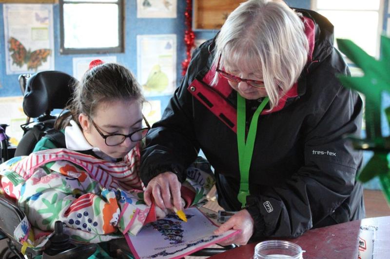 Treloar's primary student is decorating a Christmas card using a yellow crayon. her assistant is assisting her by holding the card and the crayon.