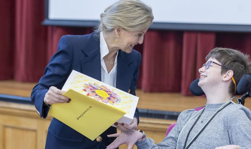 Her Royal Highness The Duchess of Edinburgh is holding a hand-made birthday card created by Treloar College student who is just next to her. Her Royal Highness and the students are both looking at each other and smiling.