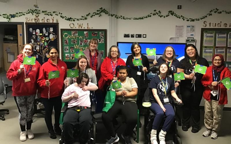 Treloar's Sing and Sign Team at Addington School posing for a group photo with staff and 2 students from their school; everyone is holding a green flag with the school's logo.