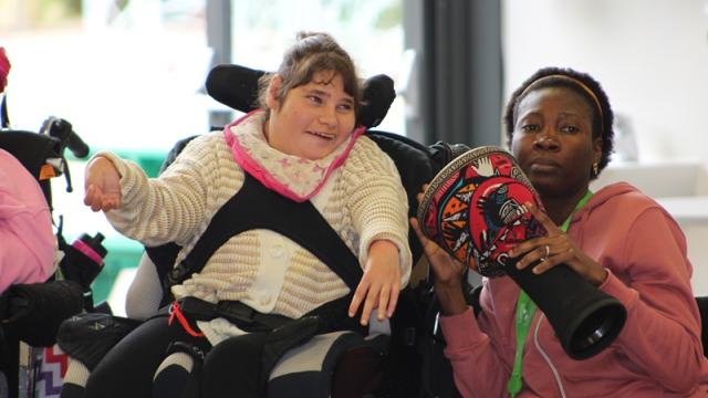 Treloar College student smiling and tapping a drum being held by her assistant during African drumming workshops.