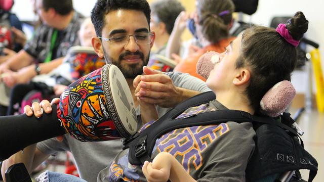 Treloar College student taking part in African drumming workshop; her assistant is holding a drum, and with his other hand he is holding the student's hand and help her tap the drum.