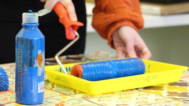 A yellow tray with blue paint in and a bottle of blue paint next to it: Rachel the art therapist is using a roller to dip in the paint.