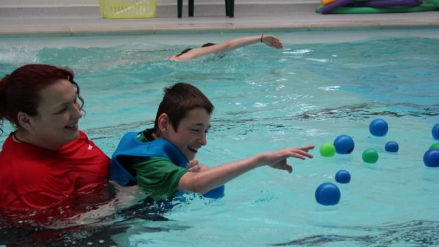 Swimming lesson: Treloar's student in the pool being supported by his assistant; the boy is trying to grab plastic balls floating on the water. Both smiling and having fun.