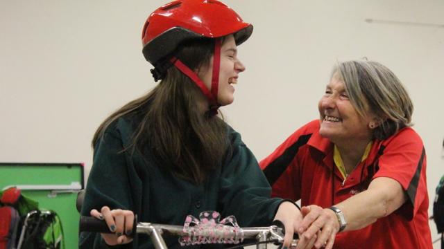 Treloar's student while triking, holding her both hands on the handlebars, wearing shiny red helmet, looking at the instructor who is helping her control the trike. Both smiling at each other.