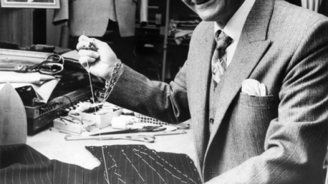 Black and white photo of Peter Little: he is sawing a suit, holding a thread and smiling at the camera.