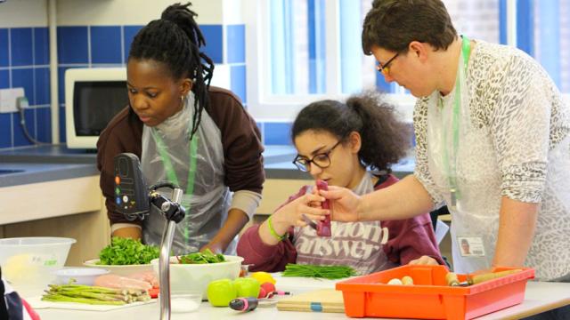 Treloar's student chopping chives with the help of her assistant. A lot of food ingredients on the table.