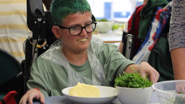 Treloar's student in the cooking classroom: bowls with food ingredients in front of him at the table and he is reaching for a bowl with chives; the boy's hair is green and he is enjoying cooking.