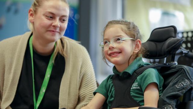 Treloar's student with her assistant in the classroom: the assistant is looking at the girl, smiling; the student is wearing glasses and looking up and smiling.