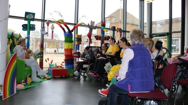 Story reading in the Jowette Centre: a group of students with their assistants sitting in semi circle, listening to a staff member reading a book.