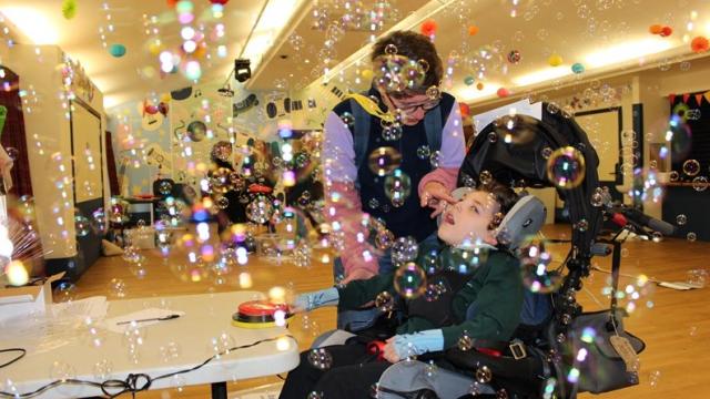 Treloar's student is using a switch to activate a bubble machine; bubbles everywhere; the parent is leaning over the student, touching his nose with a finger