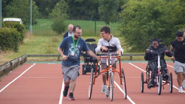 Treloar College student taking part in the annual Sports Day: he is using 3 wheeled walking frame supported by 2 members of staff.