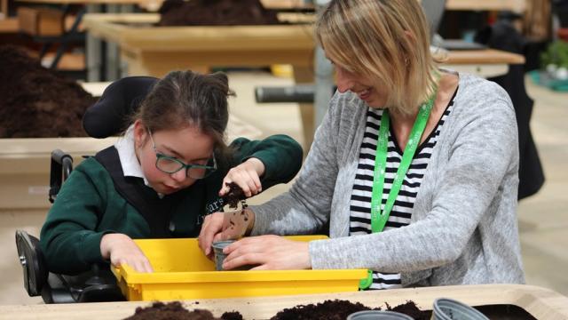 Treloar's student is using her bare hands to fill up a pot with soil; she is in the greenhouse; her assistant his holding the pot and smiling. The girl looks focused.