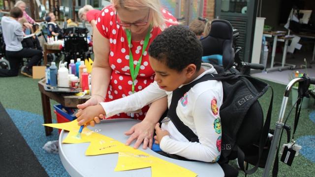 Treloar's student is decorating a yellow piece of paper that after glueing together will make a crown; his teacher is supporting him by holding his wrist and helping him put the glitter on the paper.