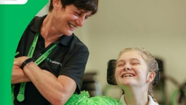 Treloar's student laughing and holding a big green pom pom; her teacher is looking at her, also laughing. Both are having a good time.