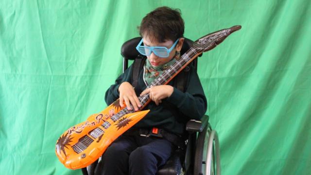 Treloar School student posing for a photo holding a guitar and wearing blue plastic sunglasses; behind him, there is a greenscreen.