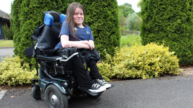 Treloar College student posing for a photo outdoors: green, tall shrubs in the background; the student is smiling shyly to the camera.