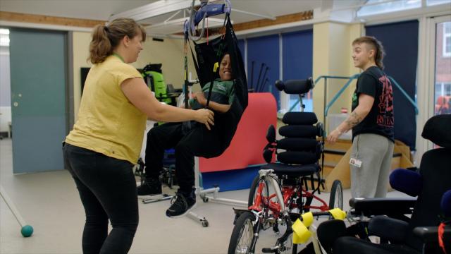 Treloar student Celeb using a hoist to access a trike with two physios assisting