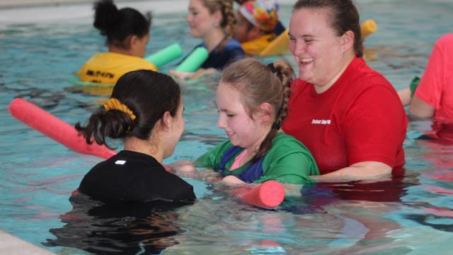 Treloar School student in the pool, supported by 2 members of staff holding onto a pink swimming noodle; in the background other students and staff.