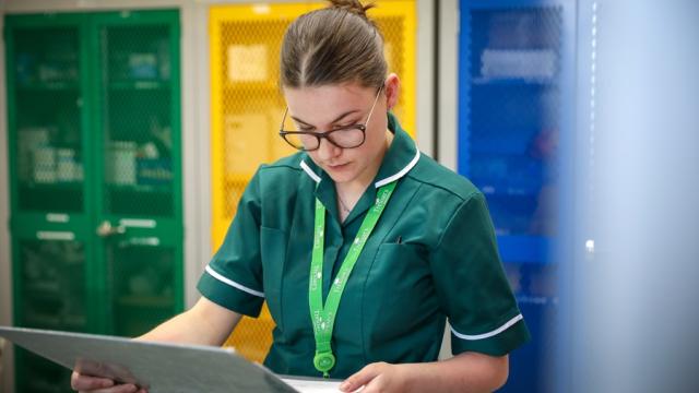 Treloar's Medicine Management Technician is holding a big medical chart in her handing and carefully looking at it; she is wearing a dark green uniform.