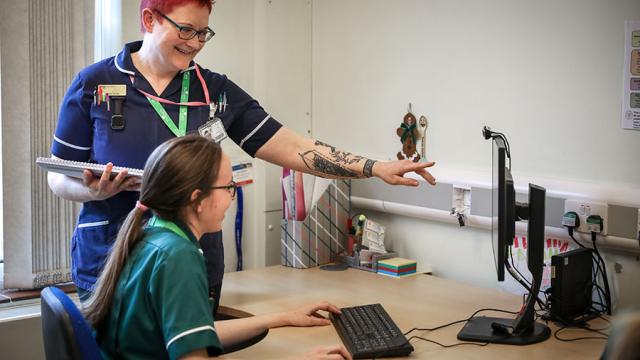 Treloar's nurse and medicine management technician in an office; the technician is sitting at her desk looking at a PC screen while the nurse is standing right next to her and pointing at the desktop, explaining something to the technician.