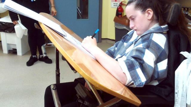 Treloar College student using a sloping desk that enables her to make notes in her book much more easily. She is in a classroom setting, she looks focused on her task.