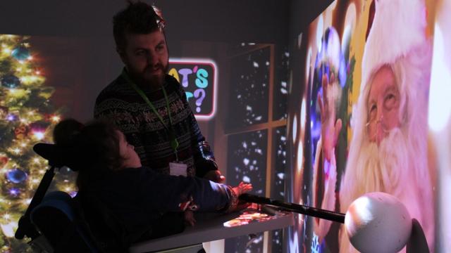 An interactive projections on the wall of Santa, accompanied by scents and sounds: Treloar College student is being assisted by her teacher to interactive with the projection by holding a big pole with a round end.