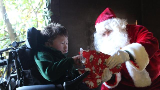 Treloar's primary student unwrapping a Christmas present gifted to him by Santa. Santa is helping him unwrap the gift wrapped in red wrapping paper.