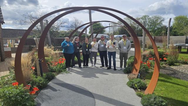 A group photo of corporate volunteers at Treloar's, all lined up n the outdoor learning area, standing underneath an archway.